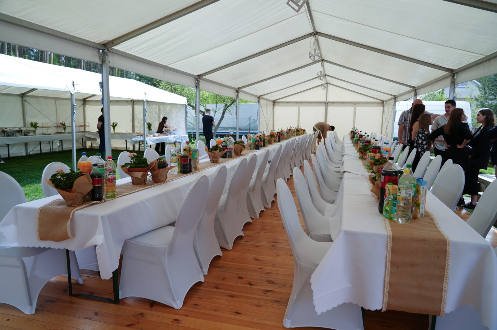 Długi stół pod białym namiotem, nakryty białym obrusem z jutowym bieżnikiem, udekorowany kwiatami w doniczkach i napojami, przygotowany na przyjęcie.