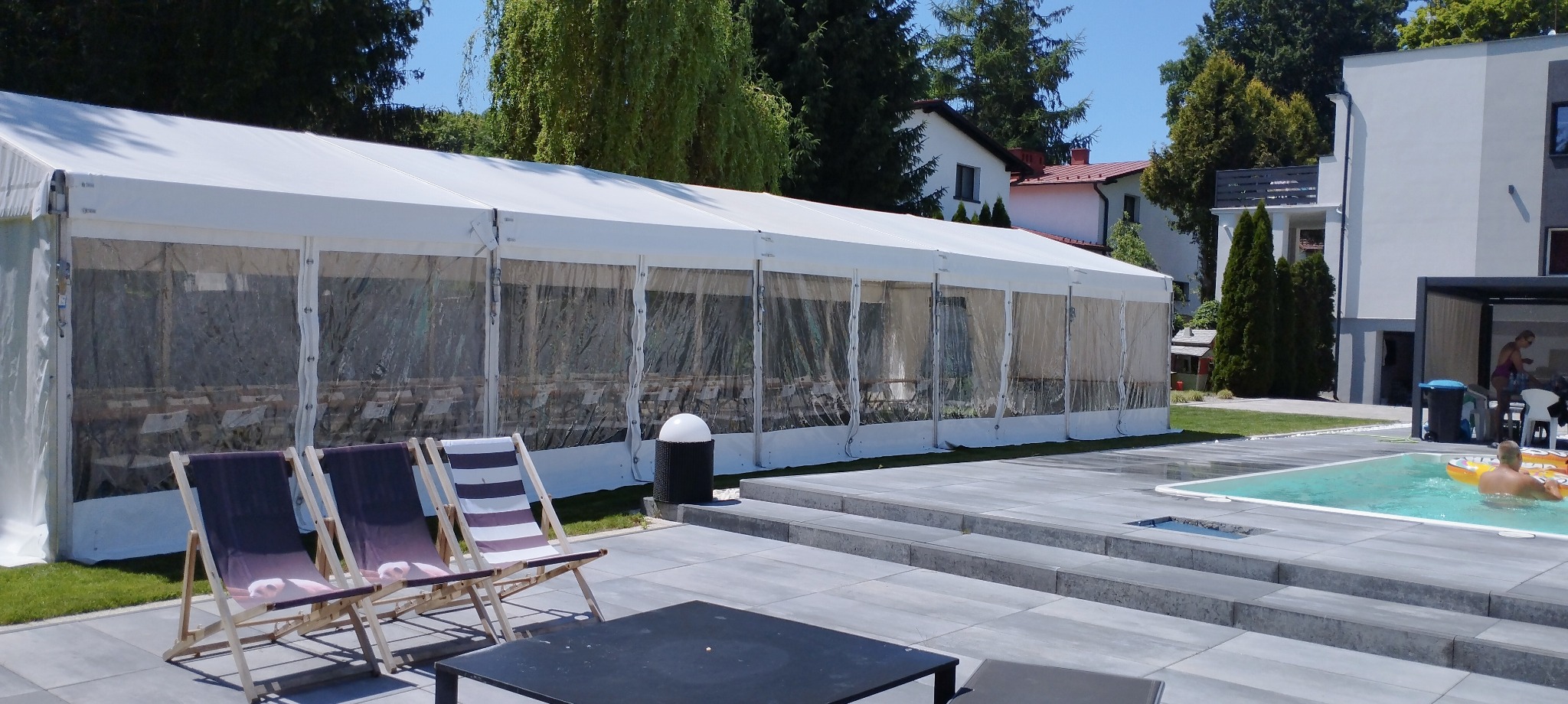 Biały namiot imprezowy z przezroczystymi ścianami, ustawiony na zewnątrz obok basenu, z widocznymi krzesłami plażowymi i stolikiem kawowym na pierwszym planie.