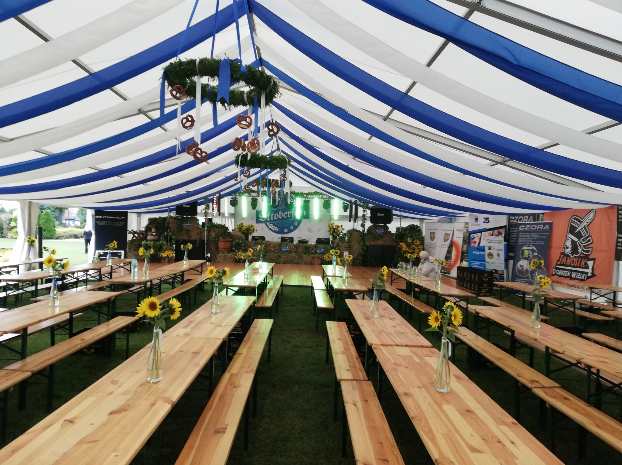 Wnętrze namiotu imprezowego z drewnianymi stołami i ławkami, dekoracjami z girland i preclami podwieszonymi pod sufitem w biało-niebieskie pasy, słoneczniki w wazonach na stołach, zielona trawa...