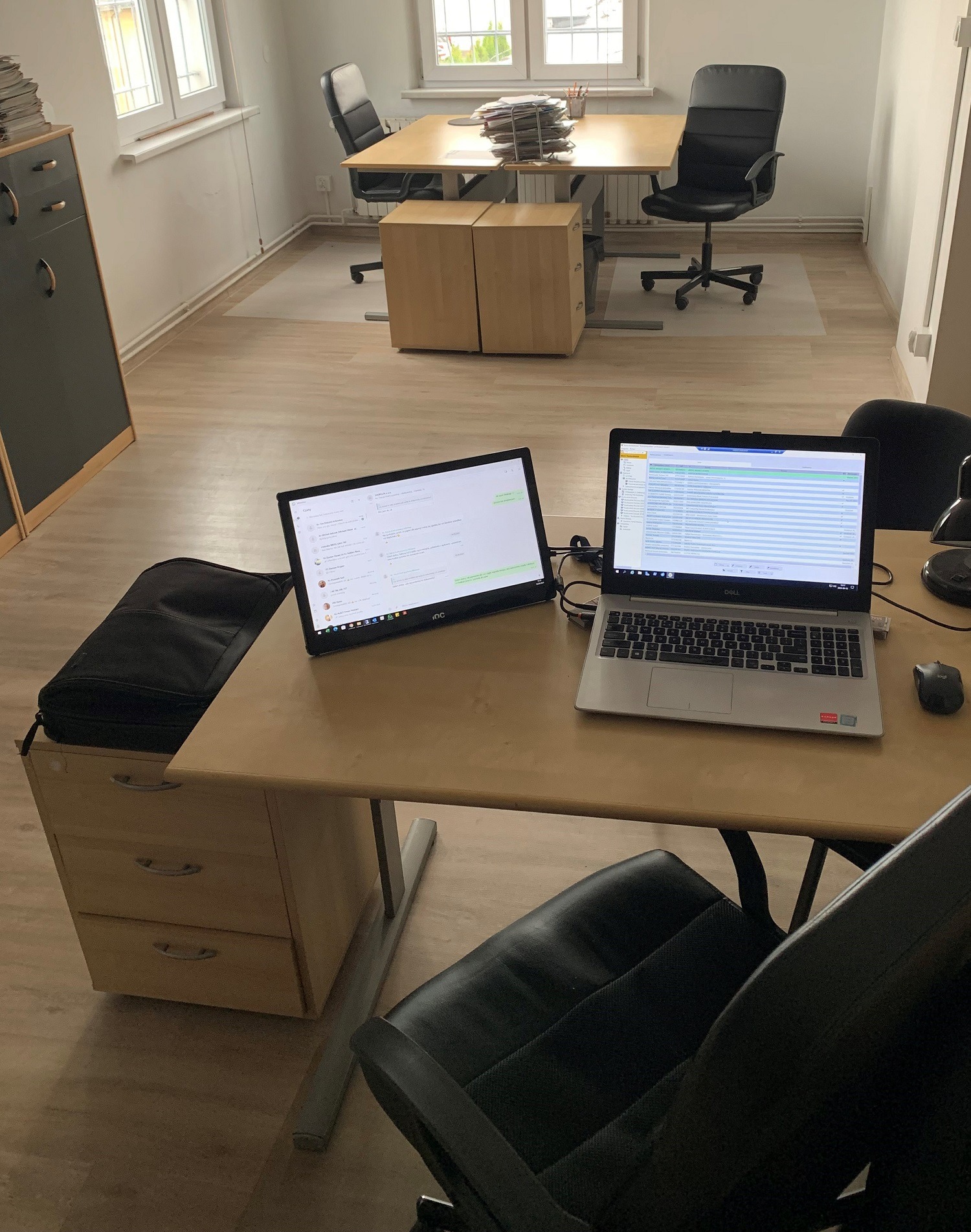 Biuro z dwoma biurkami, laptopem i dodatkowym monitorem wyświetlającym komunikator, sugerujące pracę zdalną lub hybrydową.