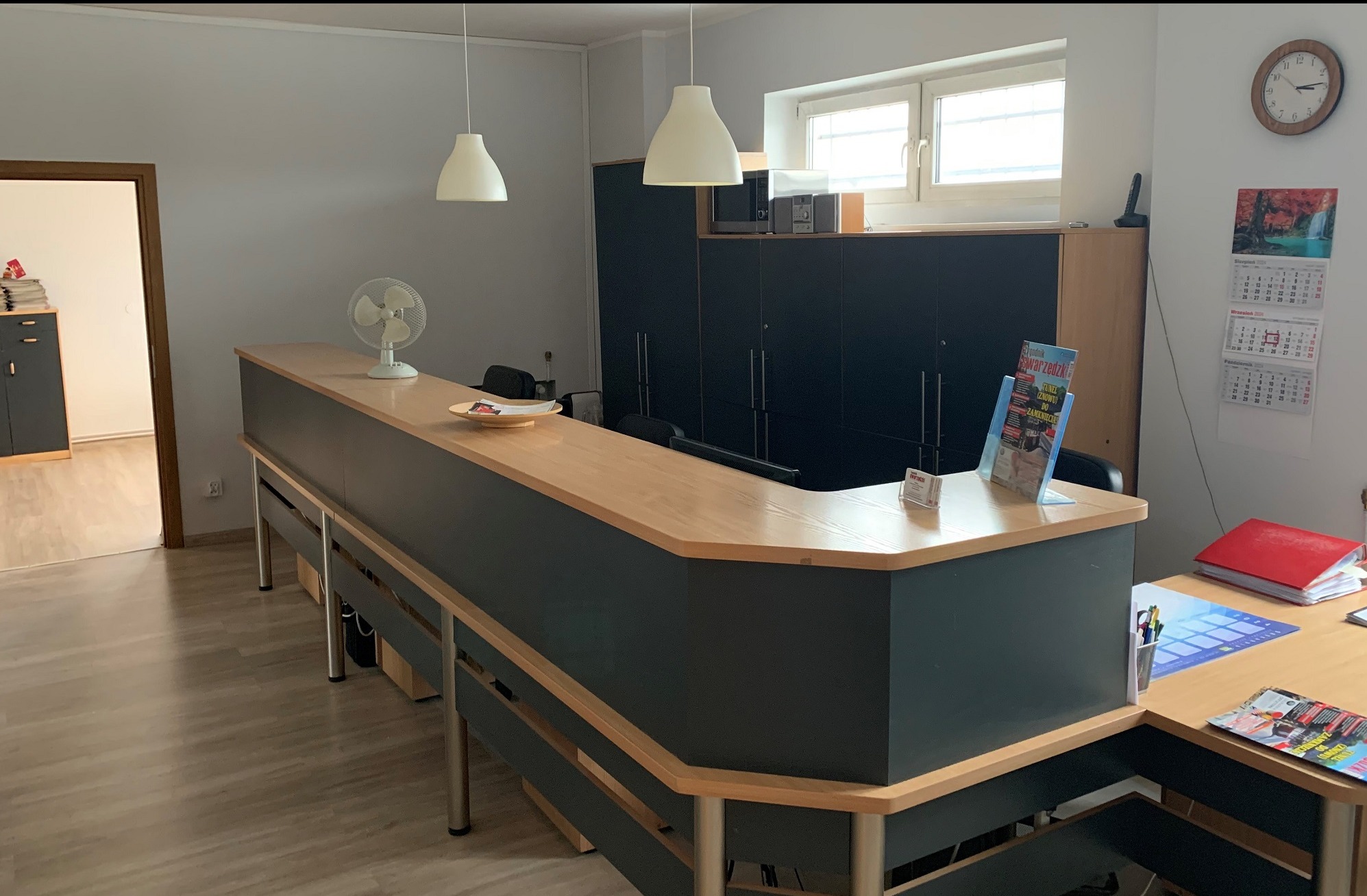 Wnętrze biura: recepcja z drewnianym blatem, wentylator, wiszące lampy, szafki w tle, kalendarz na ścianie.