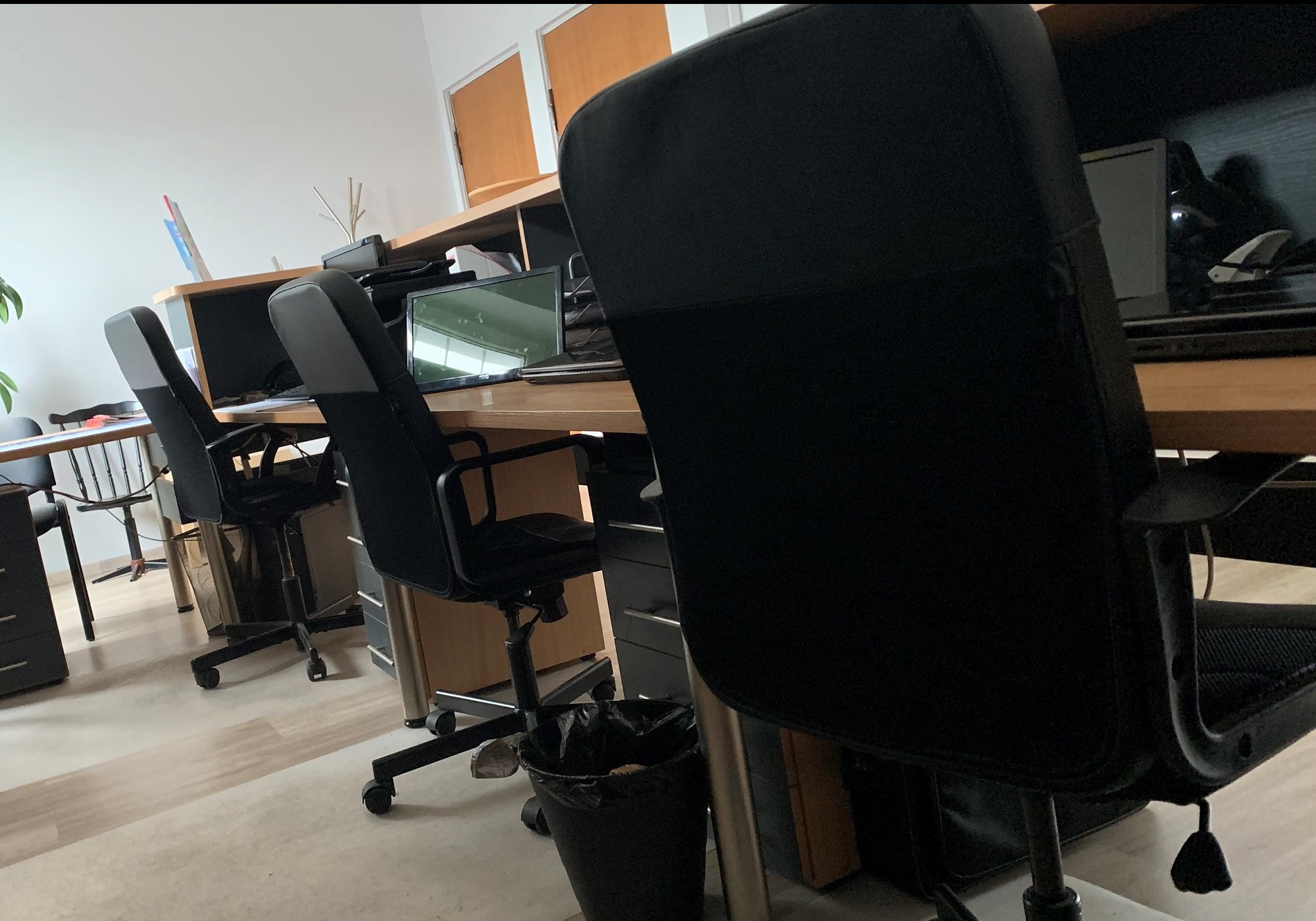 Biuro z rzędem biurek, krzesłami obrotowymi i komputerami, widok z perspektywy pracownika siedzącego przy biurku.