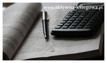 Otwarta księga z tekstem, kalkulator i pióro na blacie biurka w stonowanych kolorach.