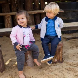 Dwoje małych dzieci, dziewczynka w różowej kurtce i chłopiec w niebieskiej koszuli, siedzą na pniakach na piaszczystym placu zabaw, prezentując modne ubrania dziecięce.