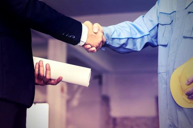 Uścisk dłoni geodety z klientem, trzymającego zwój planów, na tle surowego wnętrza budynku w trakcie budowy, z widocznym kaskiem ochronnym.