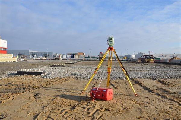 Tachimetr na statywie na placu budowy, widoczne fundamenty i walec drogowy w tle.