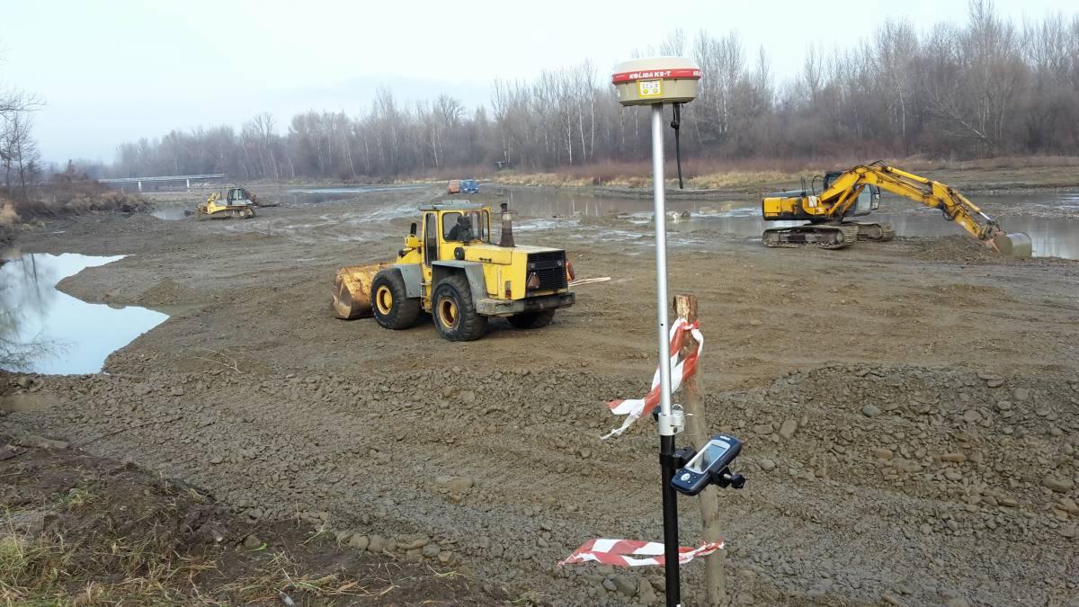 Prace ziemne w korycie rzeki: żółta ładowarka, koparka i spychacz na błotnistym terenie, w tle rzeka i drzewa, na pierwszym planie tyczka z odbiornikiem GPS.