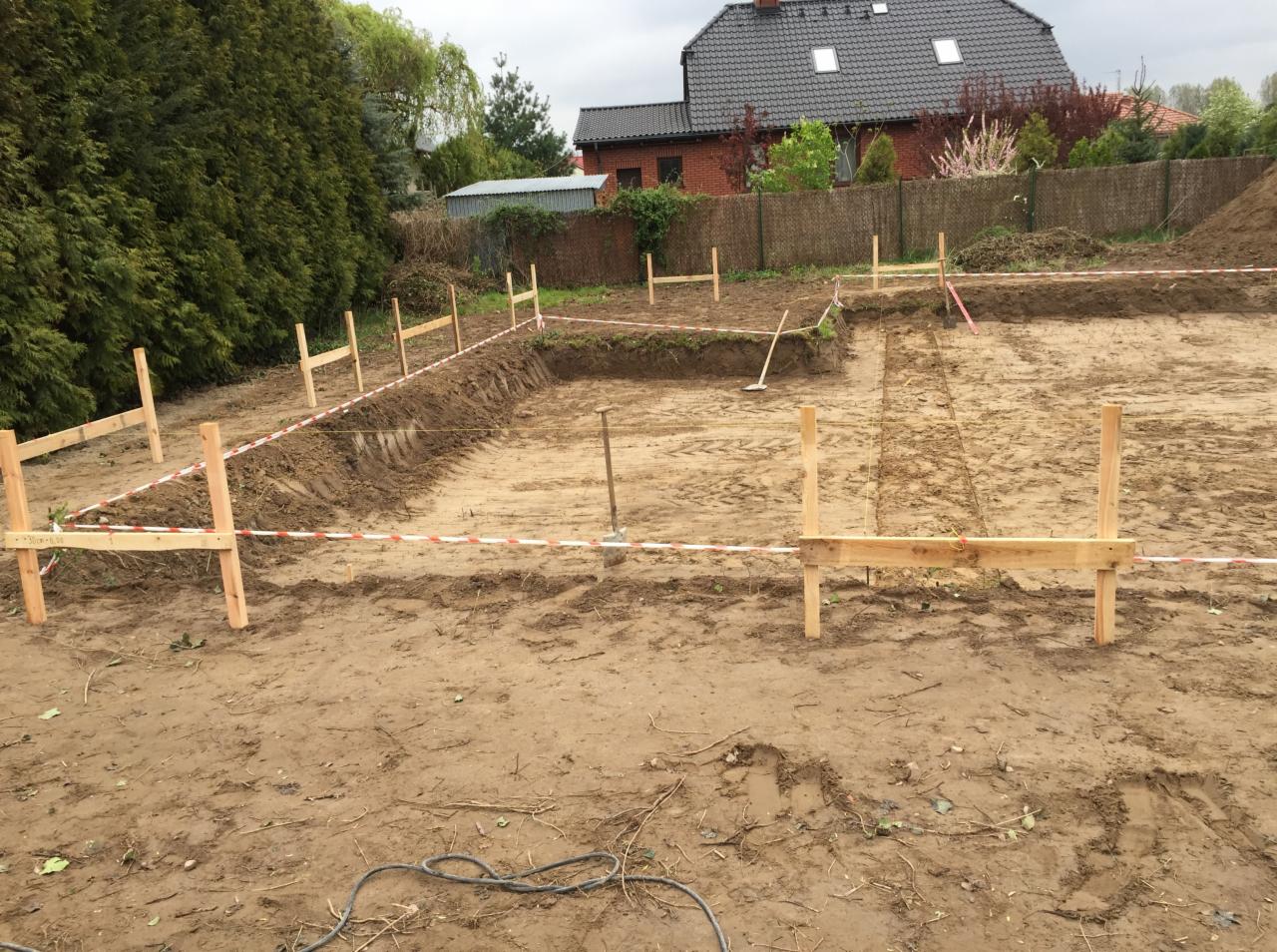 Wytyczanie fundamentów domu jednorodzinnego z użyciem drewnianych słupków, sznurka i taśmy ostrzegawczej na działce z widocznym budynkiem w tle.