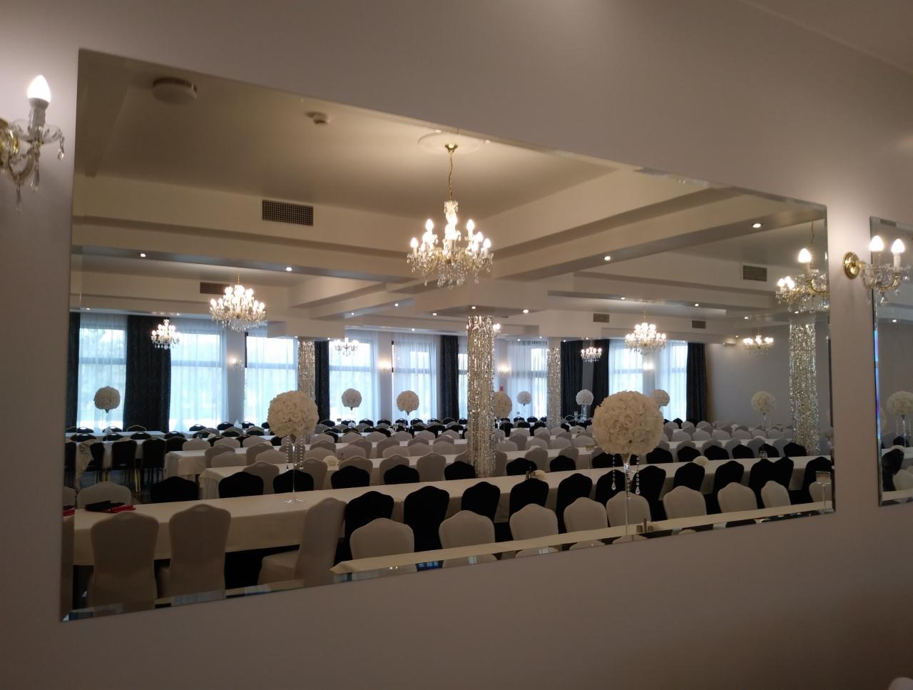 Duże lustro na ścianie odbija salę balową z białymi obrusami, czarnymi i szarymi krzesłami oraz kwiatowymi dekoracjami. Kryształowe żyrandole wiszą nad stołami. Widoczne są również kinkiety...