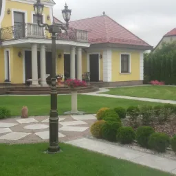 Żółty dom z czerwonym dachem otoczony zadbanym ogrodem z trawnikiem, rabatami kwiatowymi i ścieżkami z kostki brukowej, oświetlony stylową latarnią.