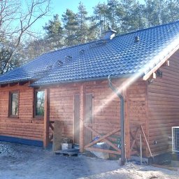 DrewLuk Łukasz Nowacki - Nowo wybudowany, parterowy dom z bali, z ciemną dachówką, widoczne orynnowanie, klimatyzator na zewnątrz i fragment lasu w tle.