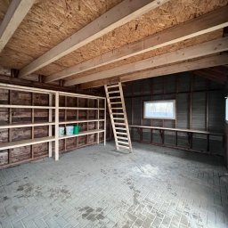 DrewLuk Łukasz Nowacki - Wnętrze szkieletowego domku gospodarczego w trakcie budowy, widoczne drewniane regały, drabina oparta o ścianę, surowy strop z desek i ceglana podłoga.