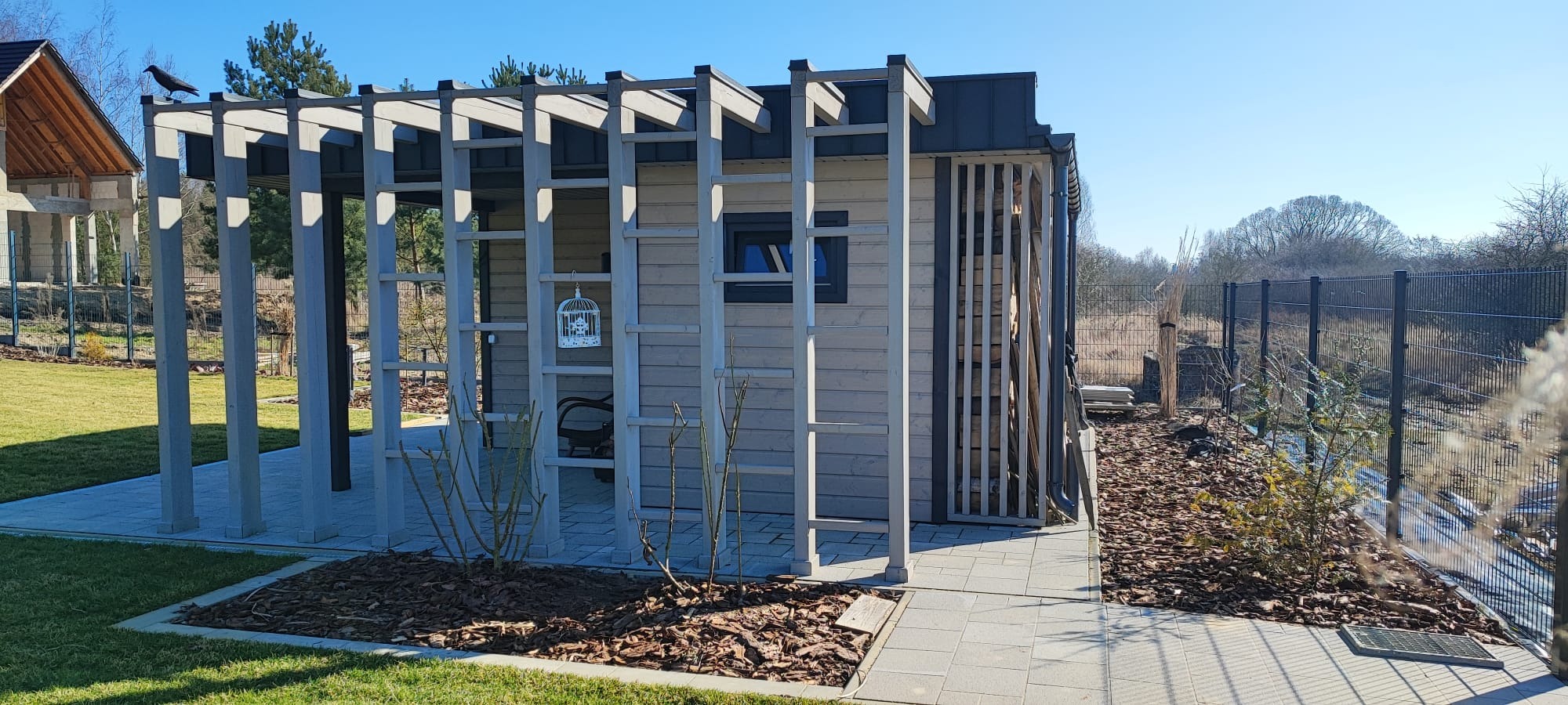 Nowoczesna drewniana altana z pergolą, widoczny kruk siedzący na konstrukcji, w tle dom w budowie.