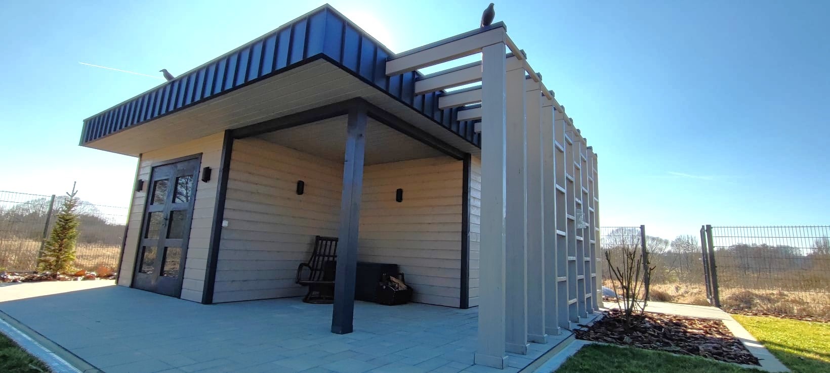 Nowoczesny, parterowy budynek z jasną elewacją, ciemnym dachem i drewnianą pergolą, na dachu siedzą dwa ptaki