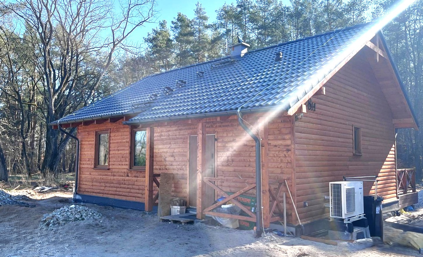 Nowo wybudowany, parterowy dom z bali, z ciemną dachówką, widoczne orynnowanie, klimatyzator na zewnątrz i fragment lasu w tle.