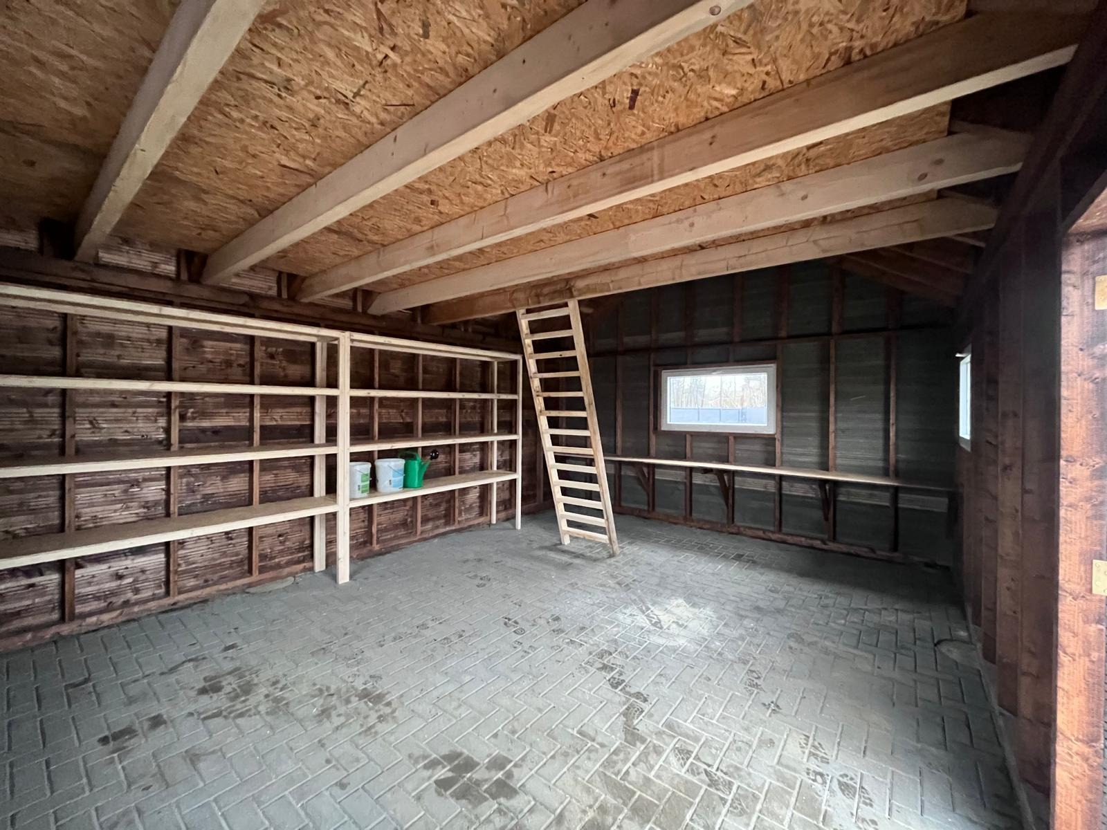 Wnętrze szkieletowego domku gospodarczego w trakcie budowy, widoczne drewniane regały, drabina oparta o ścianę, surowy strop z desek i ceglana podłoga.