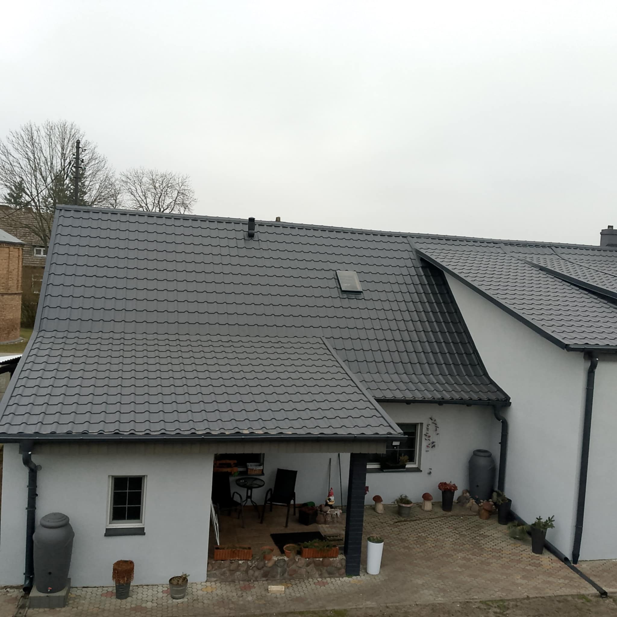 Widok z góry na gotowy, parterowy budynek z szarą dachówką, rynnami i zbiornikiem na deszczówkę, zadaszony taras z meblami ogrodowymi.