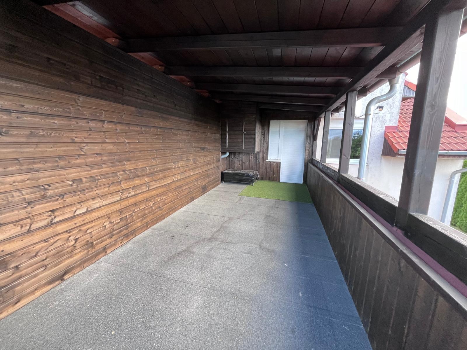 Zadaszony taras z drewnianą boazerią, szarą podłogą i fragmentem sztucznej trawy przy drzwiach.