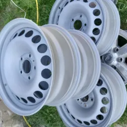 Stalowe felgi samochodowe po malowaniu proszkowym, ułożone na trawie obok drewnianego murku. Widoczne otwory wentylacyjne i srebrzysty kolor.