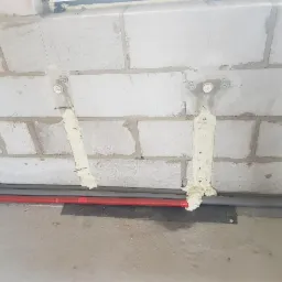 Mocowanie rur instalacyjnych do ściany z betonu komórkowego za pomocą pianki montażowej i kotew stalowych. Widoczne dwie rury: szara i czerwona.