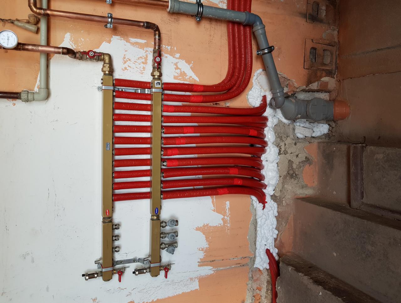 Rozdzielacz ogrzewania podłogowego z czerwonymi rurami w ścianie w trakcie montażu, widoczne elementy instalacji miedzianej i pianka montażowa.