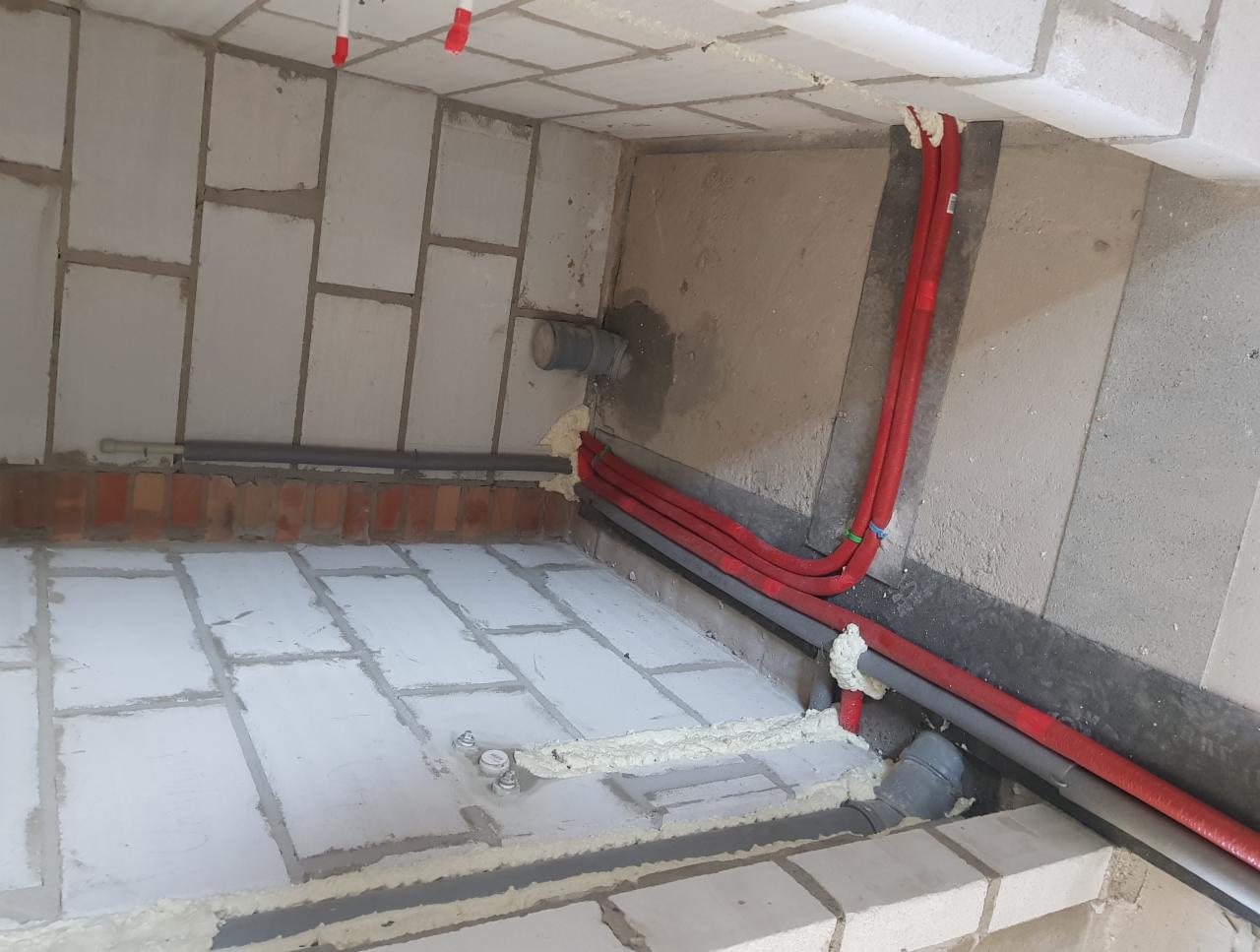 Instalacja rur w nowym domu w Grodzisku Wielkopolskim. Widoczne czerwone rury biegnące wzdłuż ściany, szare rury kanalizacyjne i białe bloczki betonowe.