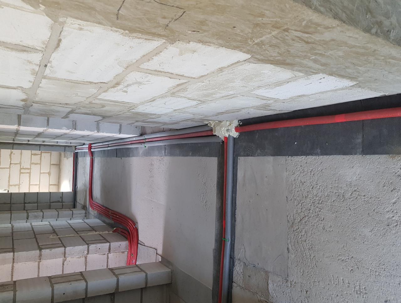 Wewnętrzne ściany w surowym stanie z widocznymi instalacjami elektrycznymi w czerwonych rurkach, częściowo zamaskowane pianką montażową, na tle betonowego stropu i schodów z bloków betonowych.