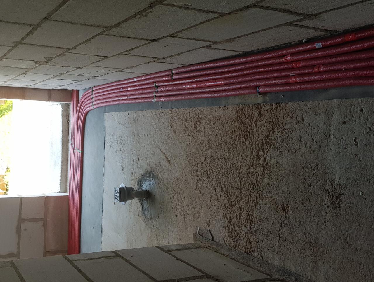 Czerwone rury instalacyjne ułożone pod sufitem w surowym stanie budynku, widoczny fragment przyłącza wodnego w podłodze.