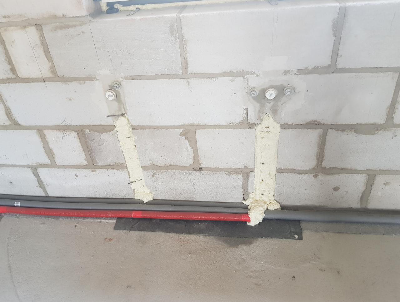 Mocowanie rur instalacyjnych do ściany z betonu komórkowego za pomocą pianki montażowej i kotew stalowych. Widoczne dwie rury: szara i czerwona.