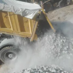 Żółta wywrotka z białym brezentem zsuwa gruz na ziemię, w tle widoczna koparka