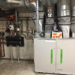 Instalacja rekuperacji Aldes z widocznymi przewodami wentylacyjnymi, rurami miedzianymi i elementami hydraulicznymi w surowym pomieszczeniu technicznym. Na urządzeniu butelka wody i pomarańczowa...