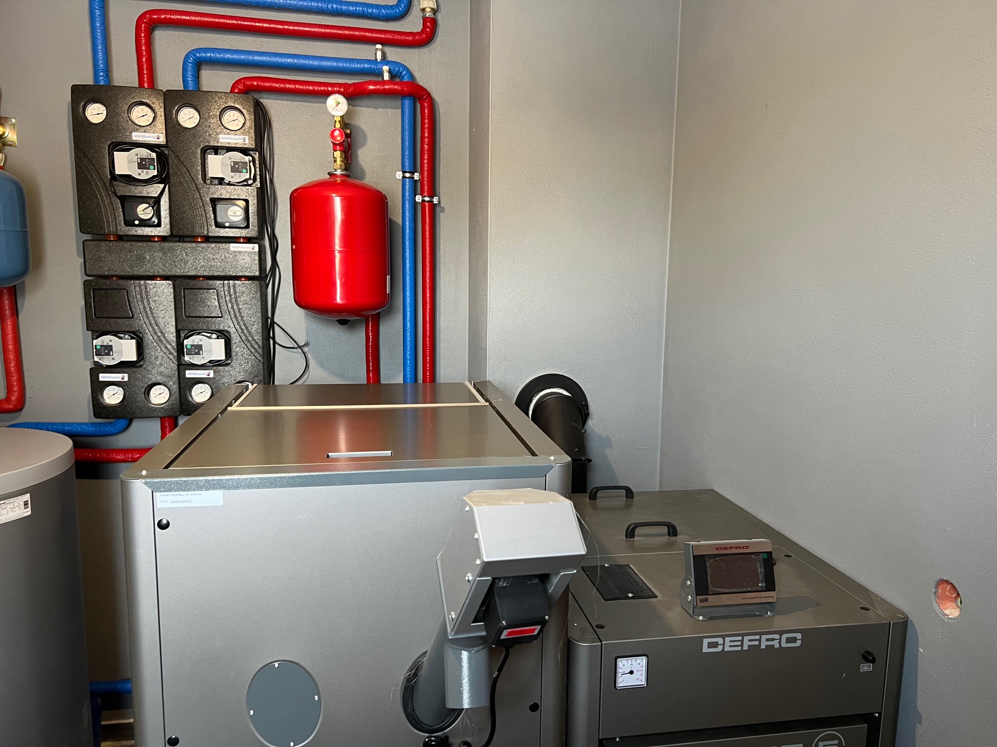 Węzeł cieplny z kotłem DEFRO, zbiornikiem wyrównawczym, czarnymi panelami sterowania i rurami w izolacji czerwonej i niebieskiej, zainstalowany w pomieszczeniu o szarych ścianach.
