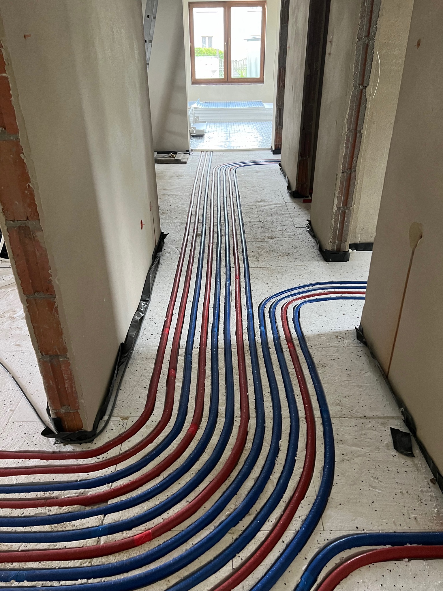 Rury ogrzewania podłogowego ułożone w jastrychu, widoczne ściany w stanie surowym, okno w tle.