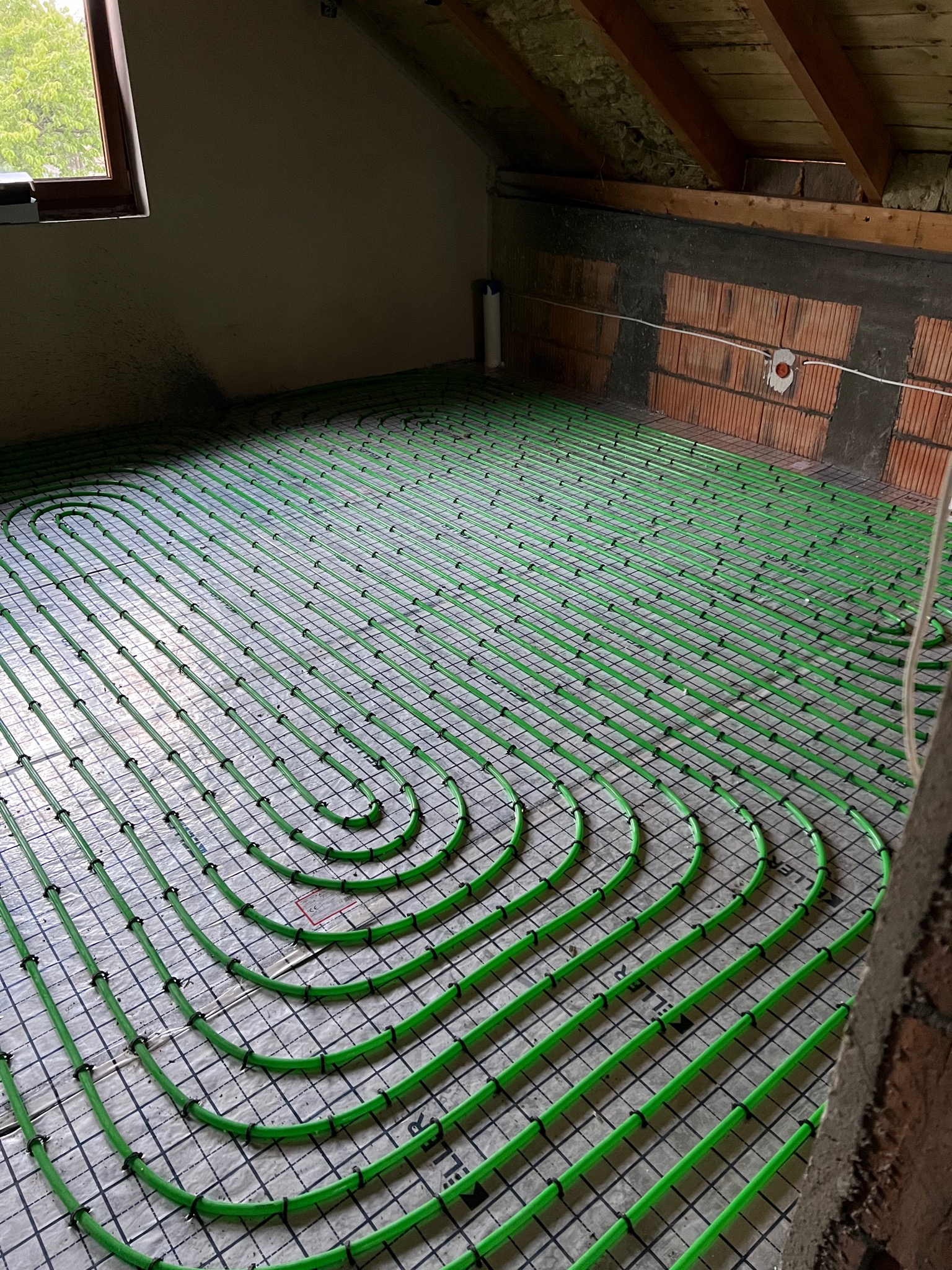 Zielone rury ogrzewania podłogowego ułożone spiralnie na izolacji z siatką w surowym wnętrzu poddasza, widoczne elementy konstrukcji dachu i fragment muru.