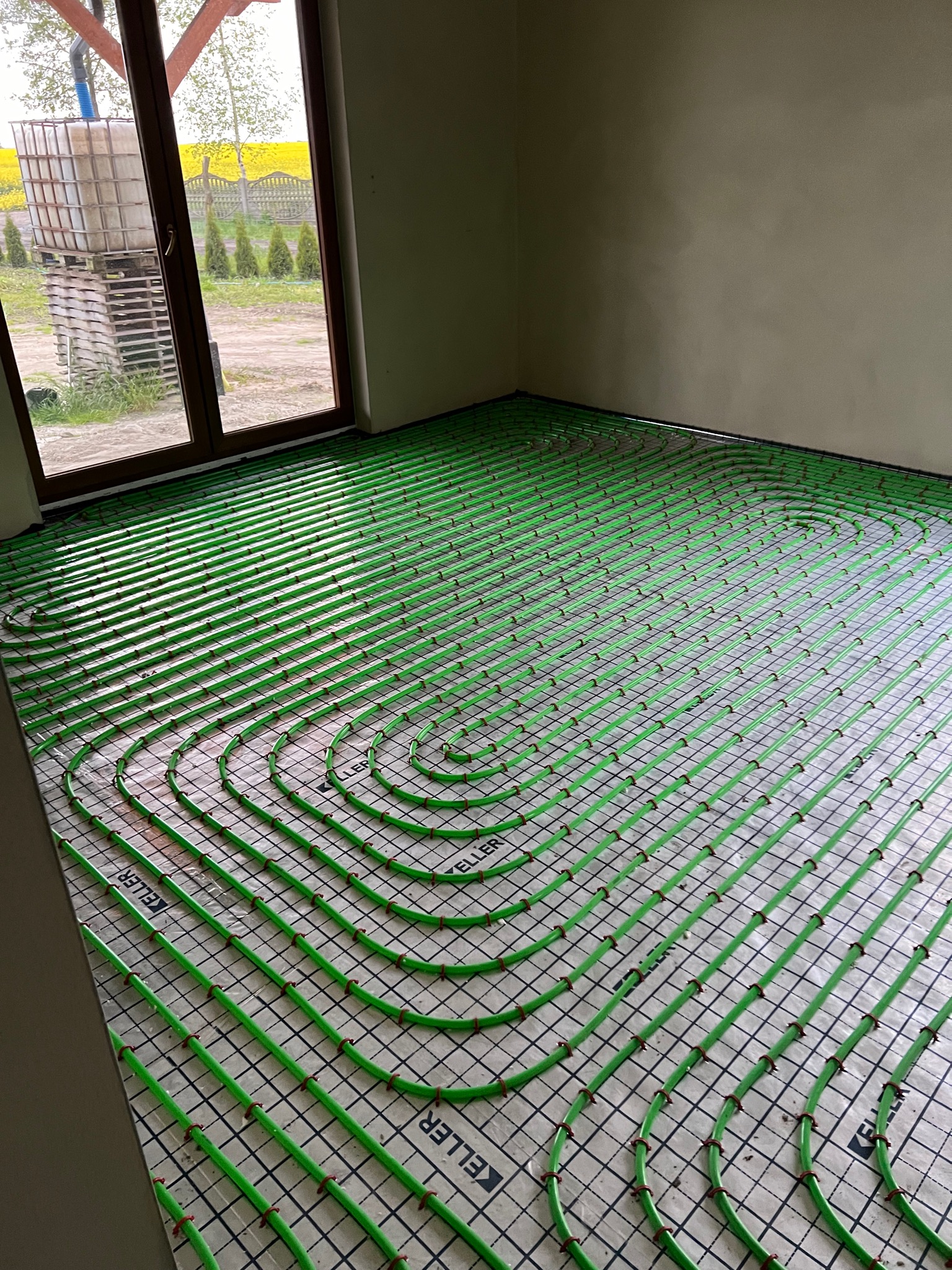 Zielone rury ogrzewania podłogowego marki Keller ułożone na folii z nadrukowaną siatką, widok na okno z widokiem na żółte pole rzepaku.