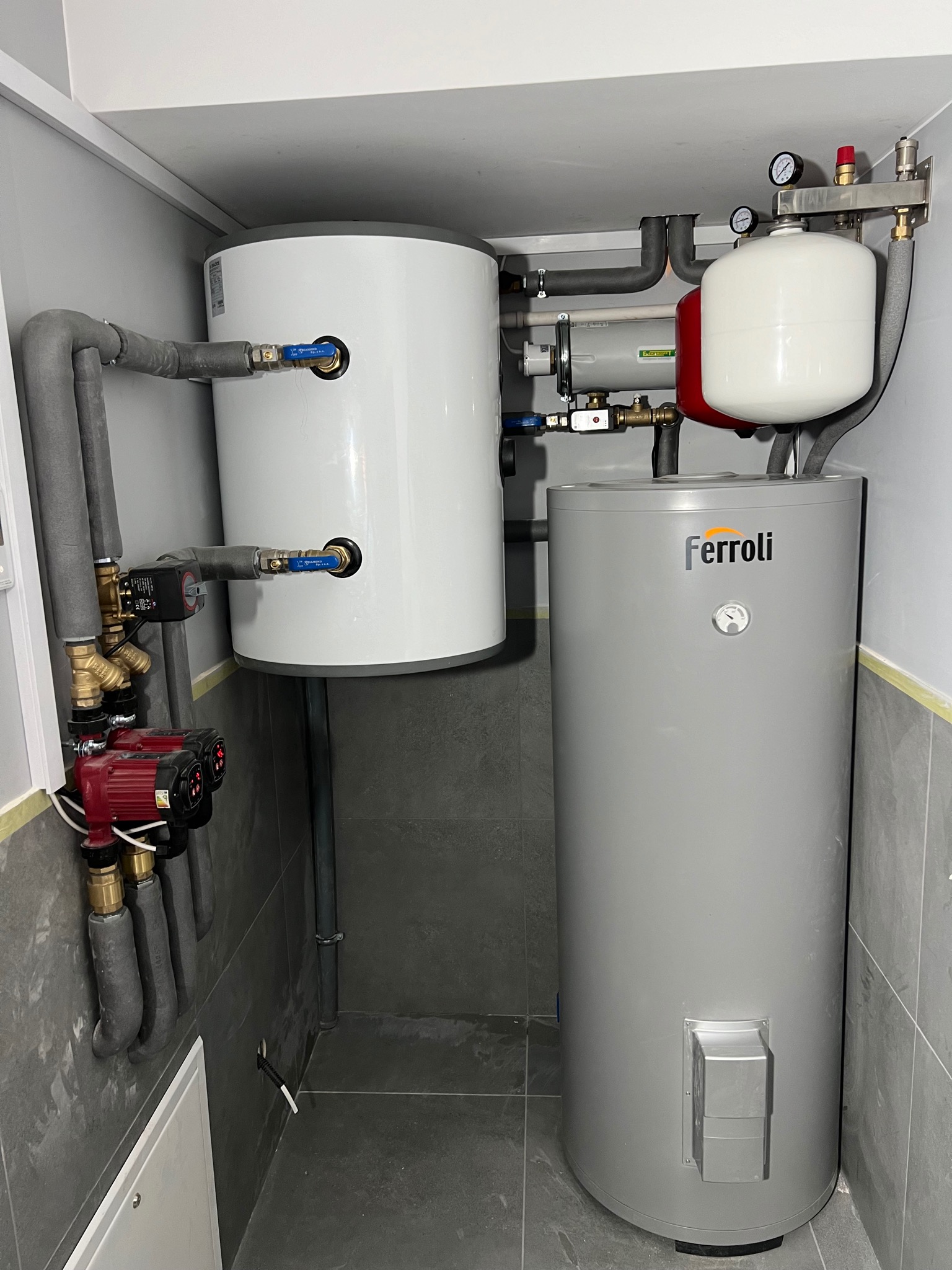 Zestaw instalacji grzewczej z dwoma zbiornikami: biały zawieszony nad szarym, widoczne pompy obiegowe, rury w izolacji oraz naczynie wzbiorcze, całość zamontowana w pomieszczeniu wyłożonym szarą...