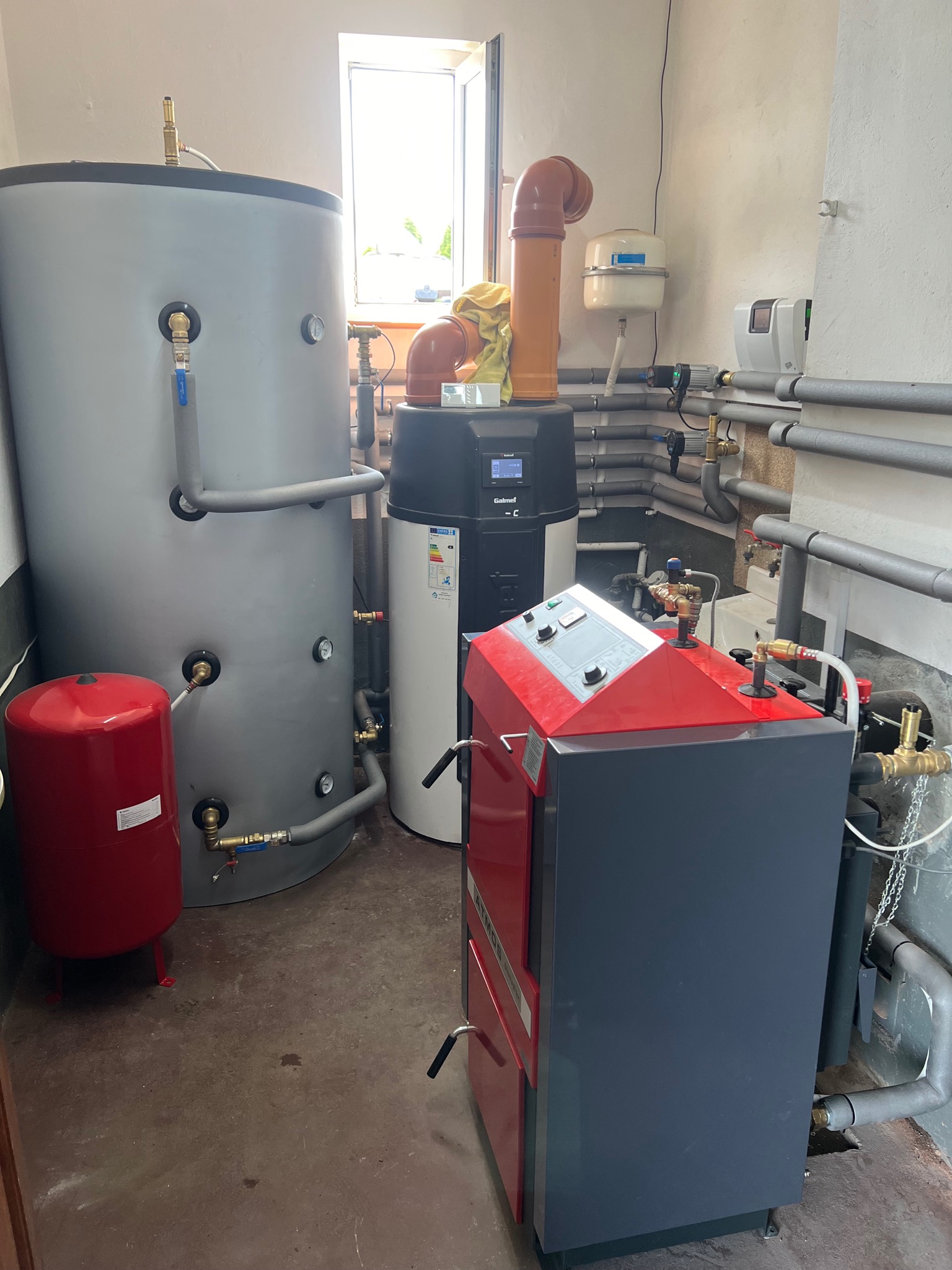 Kotłownia z kotłem na paliwo stałe z czerwonymi akcentami, zasobnikiem ciepłej wody i pompą ciepła w pomieszczeniu z szarymi rurami.