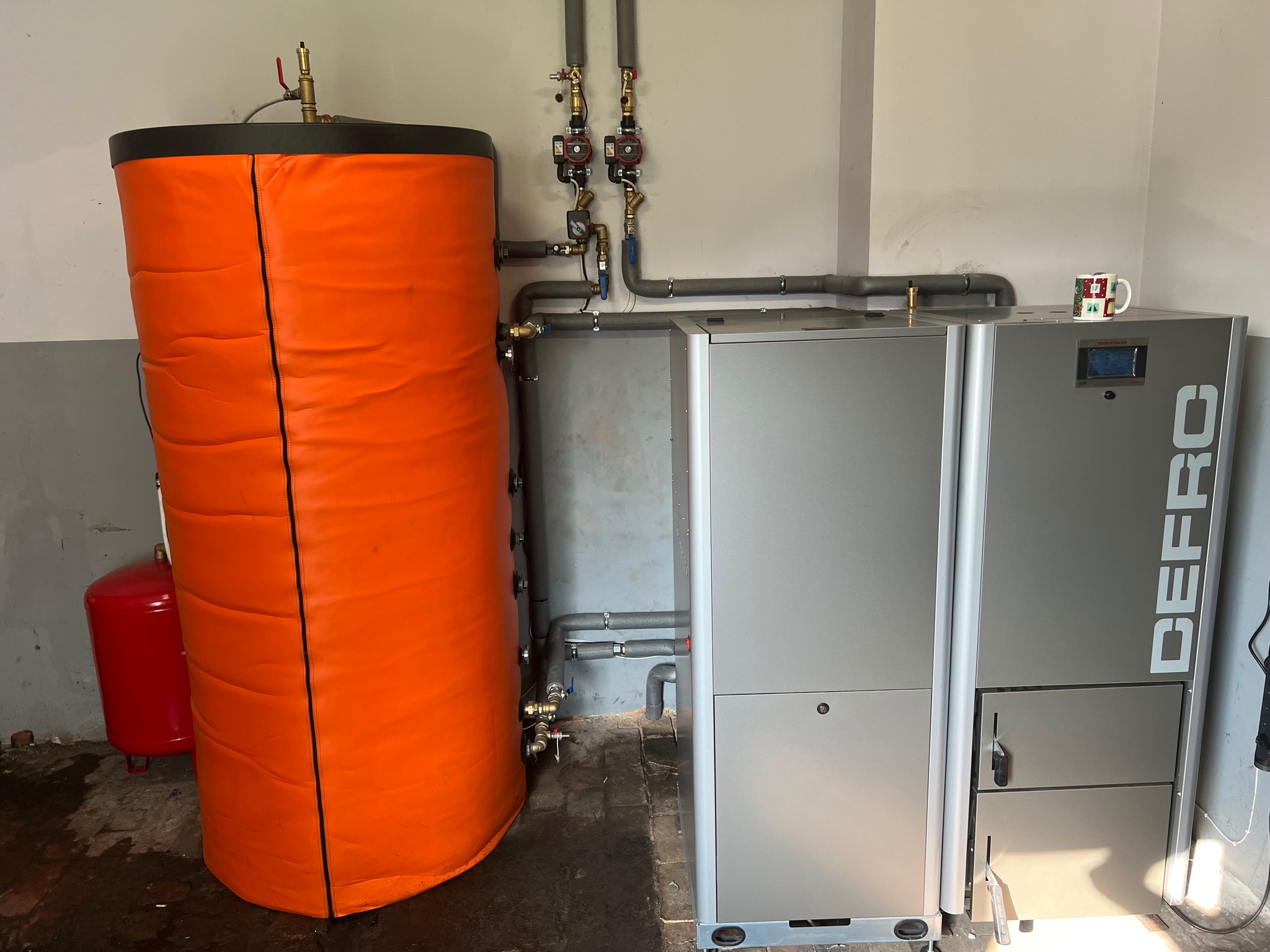 Kotłownia z piecem DEFRO, pomarańczowym zasobnikiem ciepłej wody i małym czerwonym zbiornikiem wyrównawczym, rury instalacji grzewczej w szarej izolacji, kubek z motywem świątecznym na piecu.