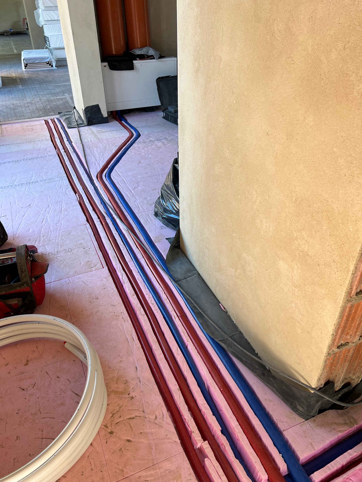 Rury grzewcze i hydrauliczne w izolacji termicznej, ułożone w wycięciach w różowej piance izolacyjnej na podłodze, widoczne narzędzia i materiały budowlane w surowym wnętrzu.
