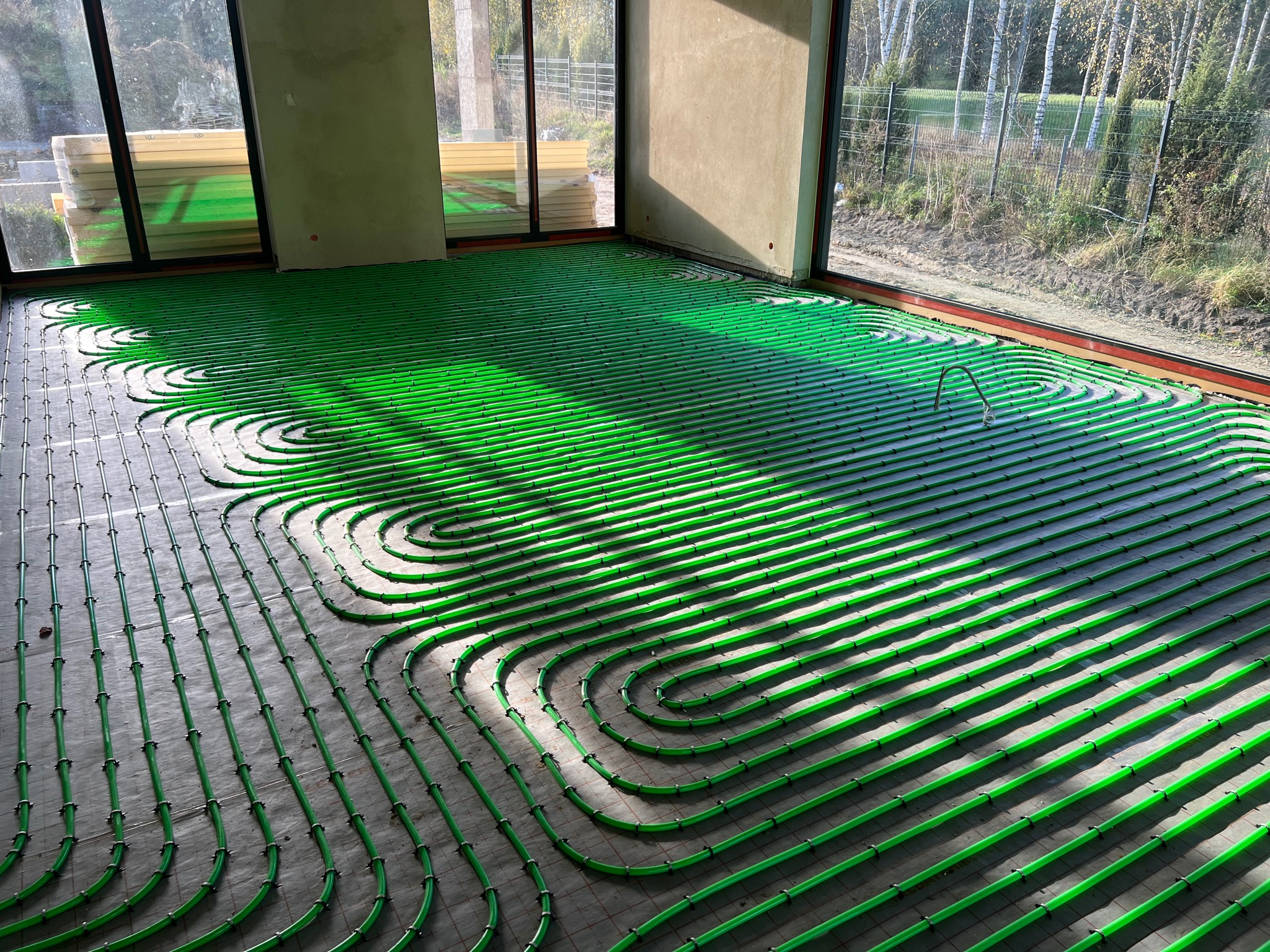 Zielone rurki ogrzewania podłogowego rozłożone w pętle na szarym podłożu w jasnym, surowym wnętrzu z dużymi oknami wychodzącymi na zewnątrz, widoczne elementy montażowe.