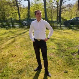 Młody mężczyzna w białej koszuli i czarnych jeansach, stojący na trawie w parku, patrzy w górę. W tle drzewa i drewniane ogrodzenie. Ujęcie w słoneczny dzień.