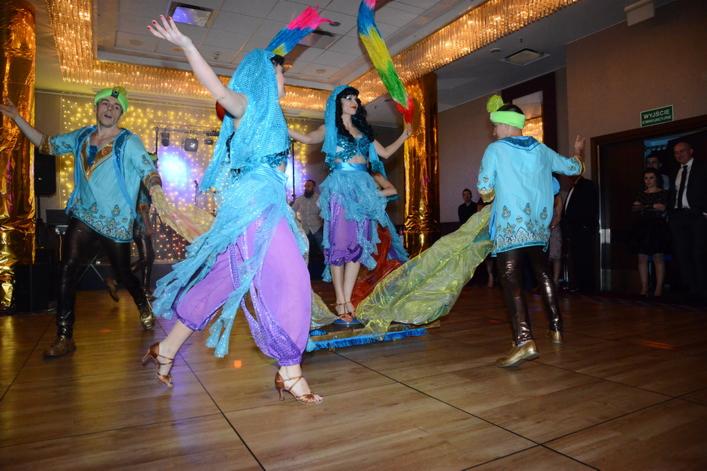 Grupa tancerzy w kostiumach inspirowanych Bliskim Wschodem, prezentujących taniec na scenie podczas wydarzenia, z widocznymi elementami dekoracyjnymi i oświetleniem.