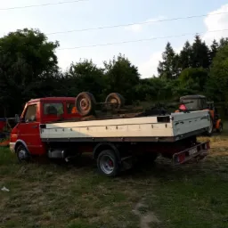 Czerwony samochód dostawczy z platformą załadowaną kołami i sprzętem, zaparkowany na trawiastym terenie w pobliżu drzew.