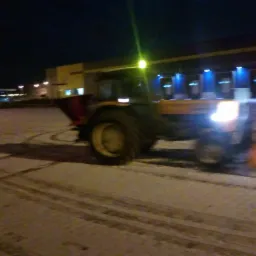 Ż&oacute;łty traktor z pługiem odśnieża oświetlony plac manewrowy w nocy, widoczne ślady opon na śniegu.