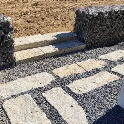 Gabiony z kamieniami, granitowe stopnie i ścieżka z nieregularnych płyt na tle drobnego żwiru.