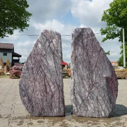 Dwa duże, pionowe kamienie z wyraźnym, białym żyłkowaniem na fioletowo-szarym tle, ustawione na kostce brukowej, z widocznymi drzewami i budynkami w tle.