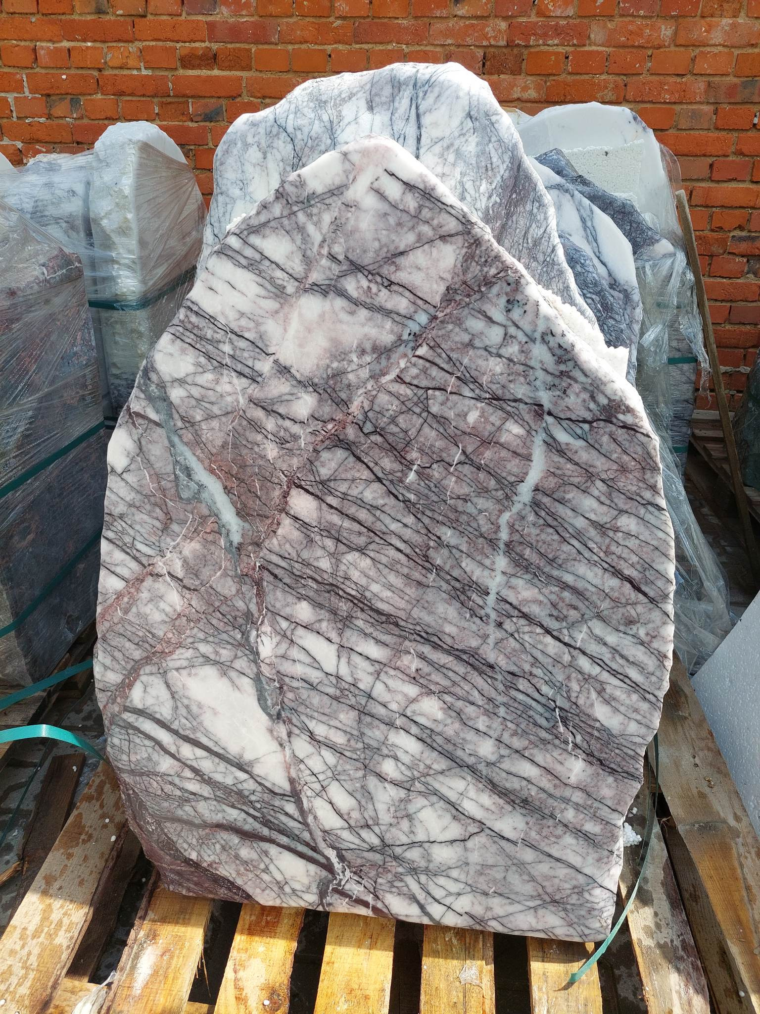 Duży, nieregularny blok marmuru z wyraźnym żyłkowaniem w odcieniach bieli, szarości i różu, ułożony na drewnianej palecie, z ceglanym murem w tle.