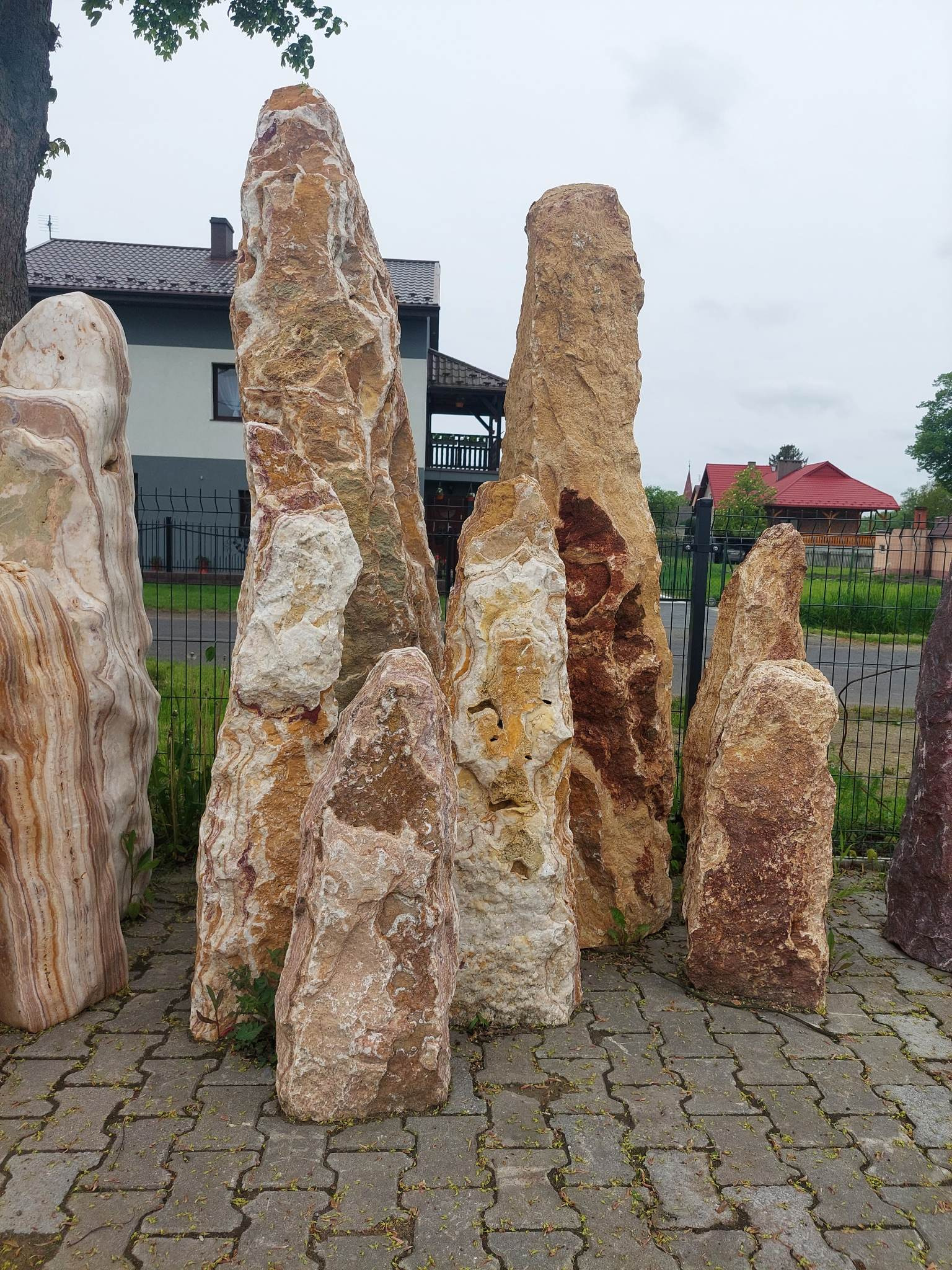 Grupa wysokich, naturalnych kamieni o zróżnicowanej kolorystyce (od beżu po brąz) ustawionych pionowo na szarej kostce brukowej, tworzących element dekoracyjny w ogrodzie lub przestrzeni publicznej...