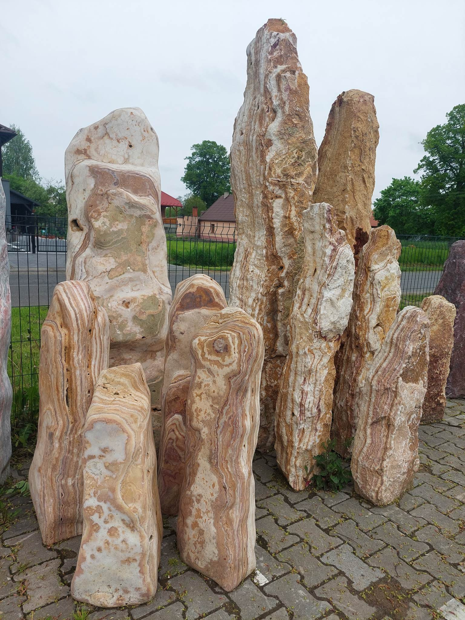 Zestaw pionowych, dekoracyjnych kamieni o warstwowej strukturze, w odcieniach beżu i brązu, ustawionych na kostce brukowej przed ogrodzeniem.