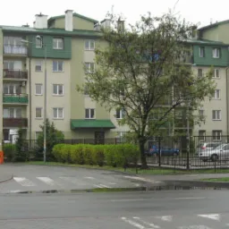 Wielopiętrowy budynek mieszkalny z balkonami, ogrodzony metalowym płotem i szlabanem, widoczny z mokrej ulicy po deszczu.